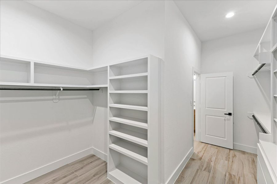 This is a spacious walk-in closet featuring ample shelving, hanging rods, and modern wood flooring. The white walls and recessed lighting provide a clean, bright atmosphere. This is a spacious walk-in closet featuring ample shelving, hanging rods, and modern wood flooring. The white walls and recessed lighting provide a clean, bright atmosphere.