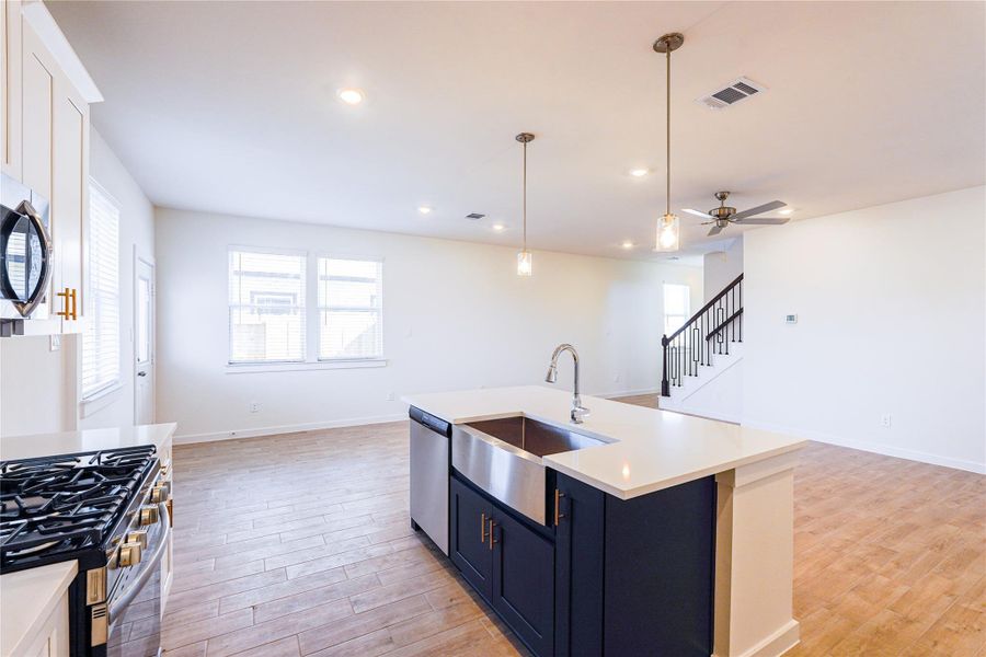 This photo showcases a spacious, open-concept kitchen and living area with modern finishes. It features a large island with a sink, pendant lighting, stainless steel appliances, and wood-style flooring. The room is bright with ample windows, and there's a staircase leading to the upper level.