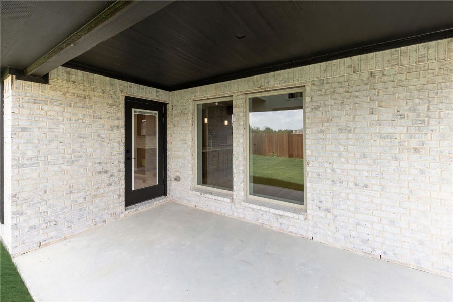 Covered back patio with concrete floor, light brick exterior, black door, and large windows overlooking fenced yard Covered back patio with concrete floor, light brick exterior, black door, and large windows overlooking fenced yard