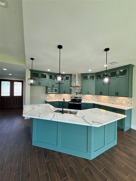 This kitchen features a modern design with teal cabinetry, a large marble island with a sink, and pendant lighting. It includes stainless steel appliances and a stylish tile backsplash, complemented by dark wood flooring. This kitchen features a modern design with teal cabinetry, a large marble island with a sink, and pendant lighting. It includes stainless steel appliances and a stylish tile backsplash, complemented by dark wood flooring.