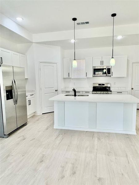 Kitchen with white cabinets, appliances with stainless steel finishes, decorative light fixtures, an island with sink, and light wood-style flooring