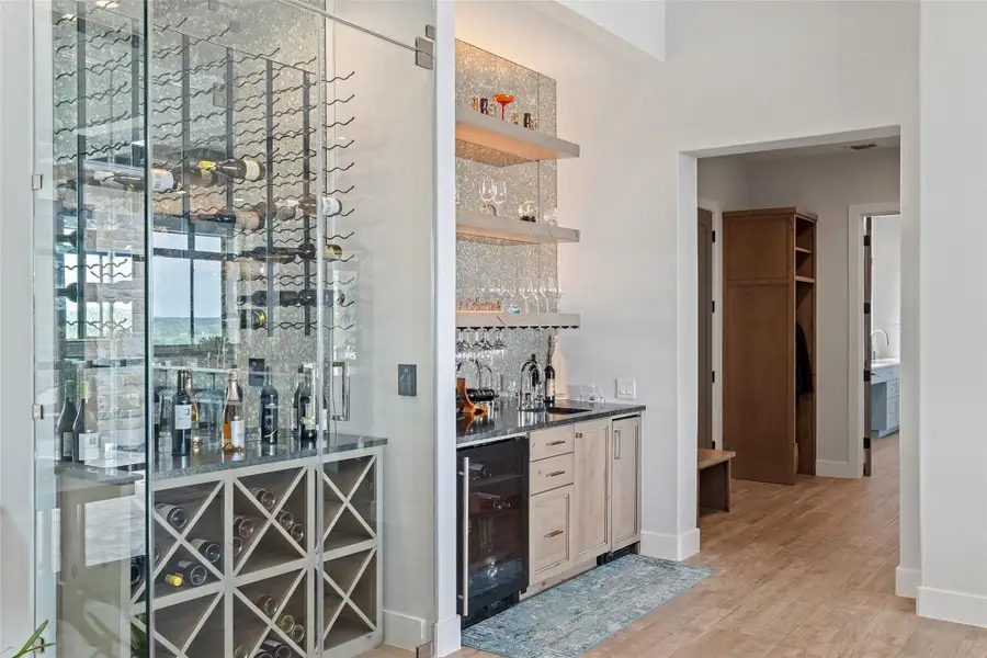 Glass-enclosed wine display with integrated bottle storage and a built-in wet bar featuring floating shelves, a beverage refrigerator, and a sink