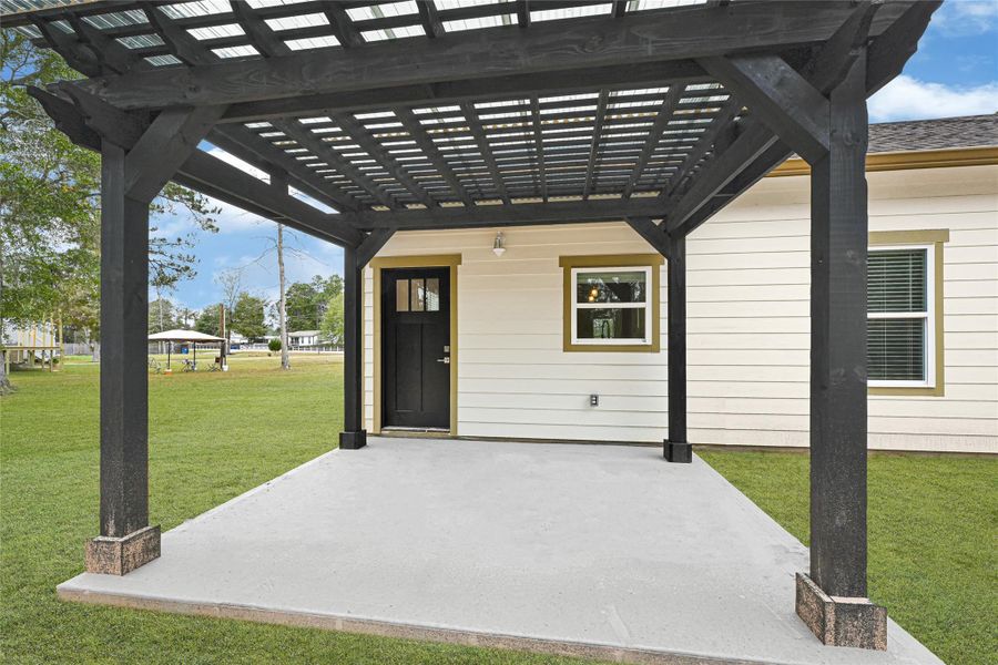 This photo shows a cozy backyard space with a modern black pergola over a concrete patio, adjacent to a light-colored house with a black door and window. The area offers a great outdoor setting for relaxation or entertaining. This photo shows a cozy backyard space with a modern black pergola over a concrete patio, adjacent to a light-colored house with a black door and window. The area offers a great outdoor setting for relaxation or entertaining.