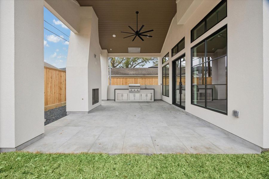 Covered outdoor living space with wood paneled ceiling, oversized ceiling fan, and built in grilling station designed for year round entertaining. Expansive black framed windows and sliding doors create seamless indoor outdoor flow, while the spacious patio layout offers room for dining and lounge seating overlooking the backyard.