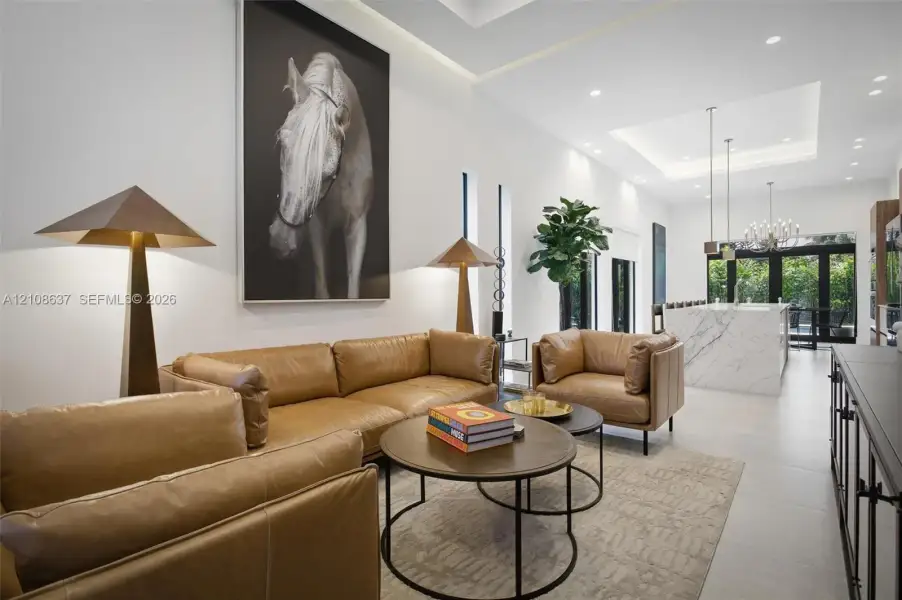 Open-concept living space featuring high ceilings with recessed tray lighting, large-format tile flooring, a kitchen island with stone-finish waterfall countertop, and multi-panel glass exterior doors