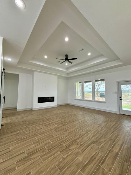 This spacious living room features a modern tray ceiling with recessed lighting and a ceiling fan. It has large windows for ample natural light, a sleek built-in fireplace, and wood-look tile flooring. A door provides access to the outdoors, enhancing the open and airy feel. This spacious living room features a modern tray ceiling with recessed lighting and a ceiling fan. It has large windows for ample natural light, a sleek built-in fireplace, and wood-look tile flooring. A door provides access to the outdoors, enhancing the open and airy feel.