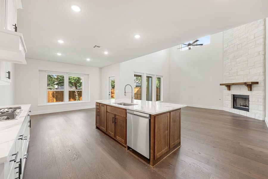 Open kitchen with wood island, quartz countertop, and stainless dishwasher, open to living area with stone fireplace and hardwood floors