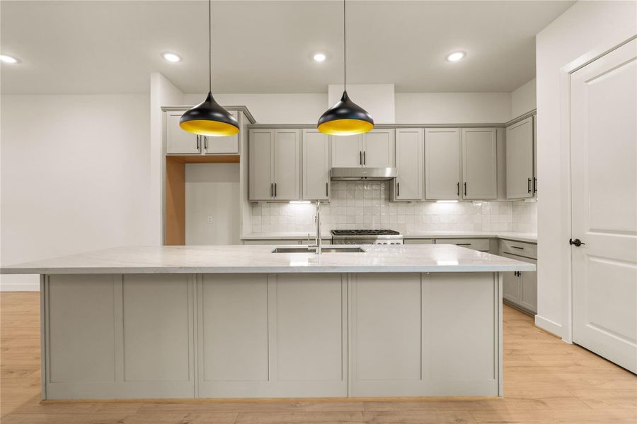 The culinary center — expansive quartz prep island with undermount double sink, stainless steel range hood, under-cabinet lighting, and clean tile backsplash.