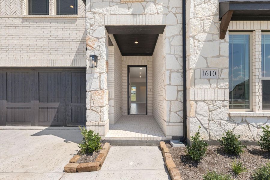 Front entry of home with white brick and stone exterior, dark wood garage door, and covered doorway with wall lantern