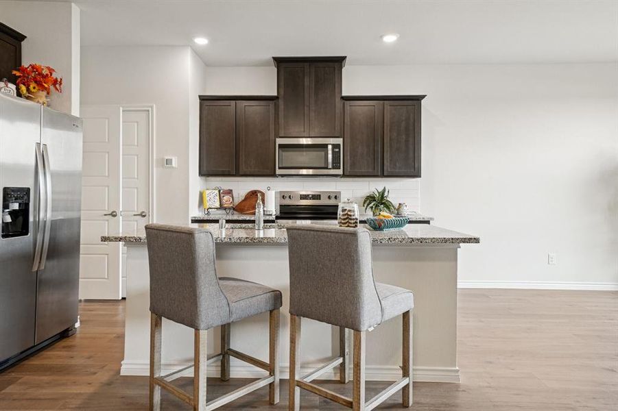 Kitchen with dark brown cabinetry, an island with sink, light stone counters, appliances with stainless steel finishes, and backsplash Kitchen with dark brown cabinetry, an island with sink, light stone counters, appliances with stainless steel finishes, and backsplash