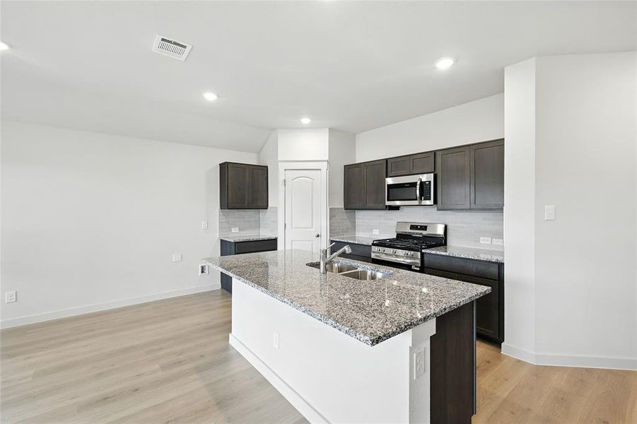 Kitchen featuring stainless steel appliances, light stone countertops, backsplash, a kitchen island with sink, and light wood finished floors Kitchen featuring stainless steel appliances, light stone countertops, backsplash, a kitchen island with sink, and light wood finished floors