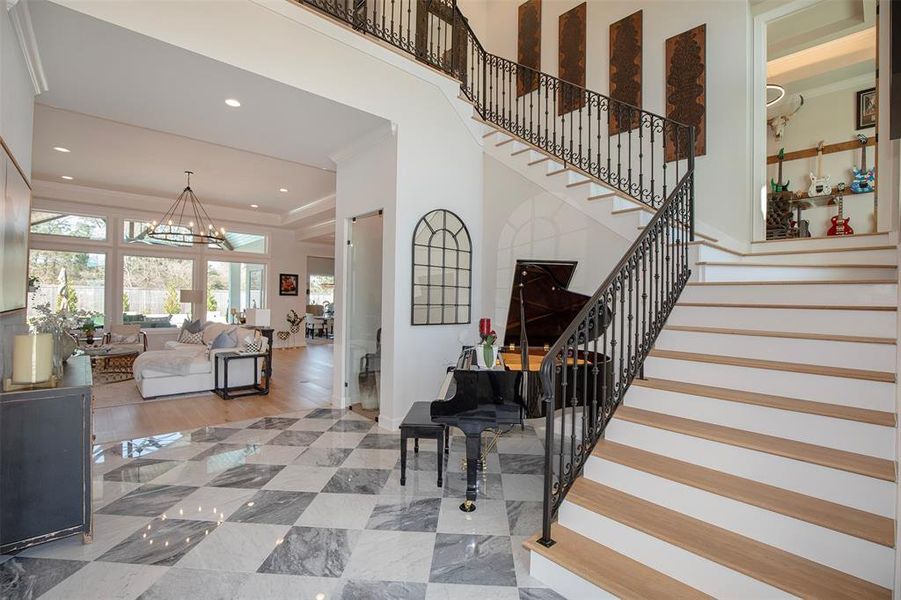 This elegant home features a spacious, open living area with large windows providing ample natural light. A grand staircase with ornate railings leads to the upper level. The entryway includes polished marble flooring and a stylish piano, enhancing the sophisticated ambiance.