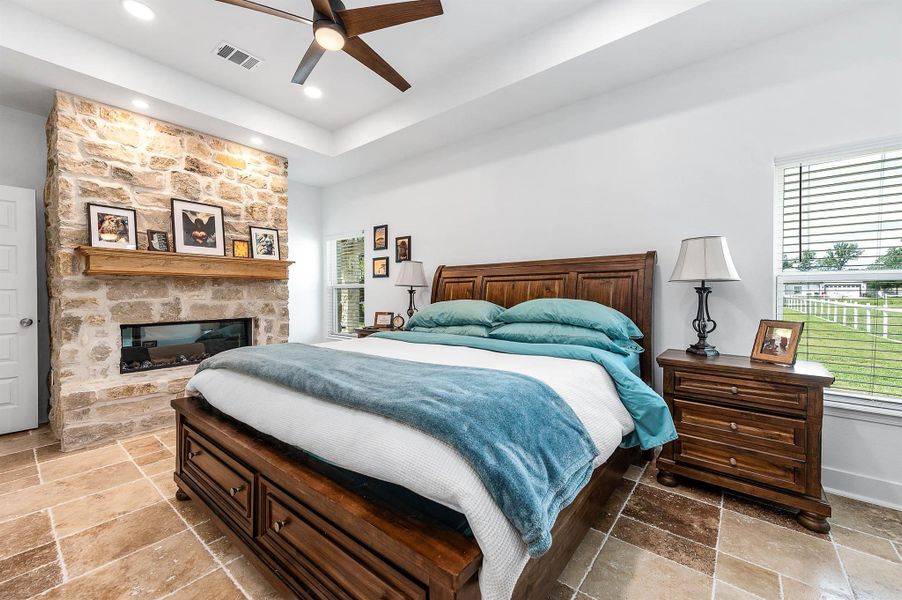 This inviting bedroom features a beautiful stone fireplace with a rustic wood mantel, creating a warm and cozy focal point. Large windows provide natural light, while the thoughtful finishes and spacious layout offer comfort and charm throughout.