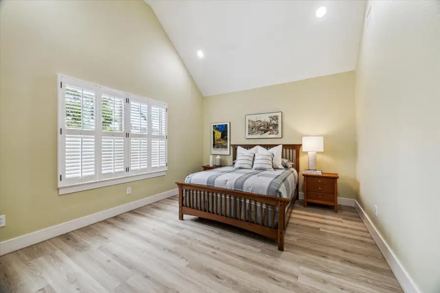 The primary bedroom features a vaulted ceiling, neutral walls, wood flooring and recessed lighting. The view overlooks the backyard patio and greenspace.