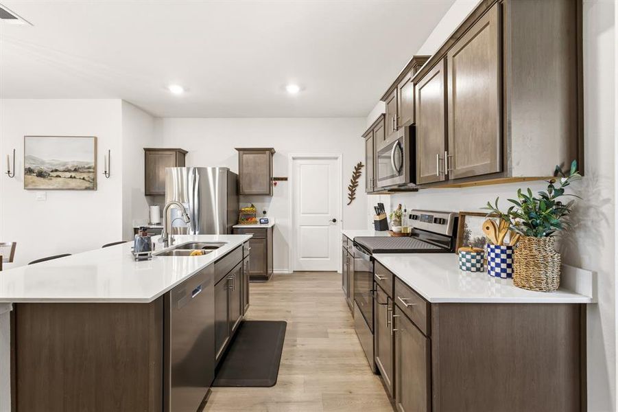 The kitchen features a large island with modern appliances, white countertops, tons of dark wood cabinetry, and stainless-steel appliances The kitchen features a large island with modern appliances, white countertops, tons of dark wood cabinetry, and stainless-steel appliances