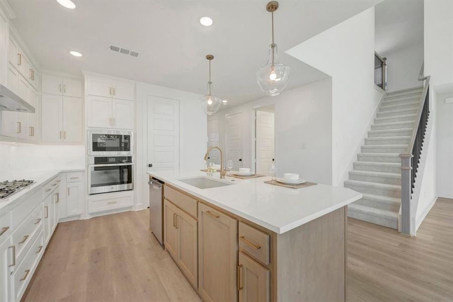 Bright kitchen featuring white cabinetry, light countertops, a central island with dual pendant lighting, stainless steel double wall ovens, and wood-finish flooring extending to a carpeted staircase with a dark railing