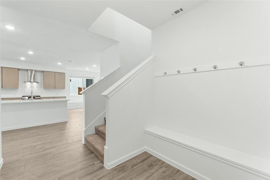 Mudroom with built-in bench and hooks near staircase, open to kitchen with tan cabinets and range hood