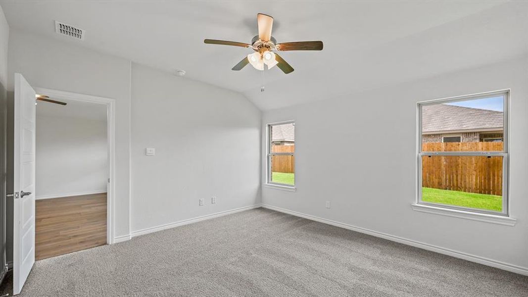 Spacious room featuring vaulted ceilings, a ceiling fan with light fixture, gray carpet flooring, two windows, and an open doorway revealing wood-finish flooring Spacious room featuring vaulted ceilings, a ceiling fan with light fixture, gray carpet flooring, two windows, and an open doorway revealing wood-finish flooring