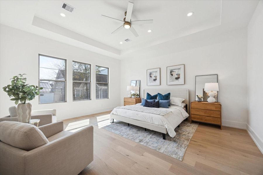 Light-filled primary suite showcasing a tray ceiling, three large windows, recessed lighting, and space to create a relaxing sitting area.