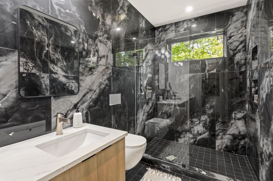 Bathroom featuring floor-to-ceiling dark marble-patterned tile, a wood-finish vanity with a white countertop and integrated sink, a glass-enclosed shower with dark tile flooring, a horizontal window, and a wall-mounted rectangular mirror