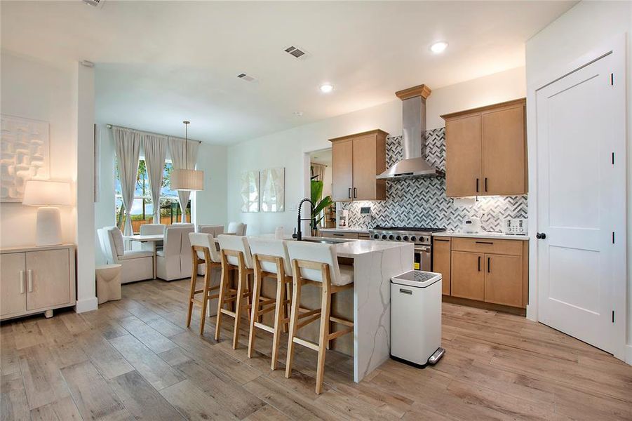 Kitchen featuring wall chimney range hood, backsplash, a center island with sink, light stone countertops, and a breakfast bar Kitchen featuring wall chimney range hood, backsplash, a center island with sink, light stone countertops, and a breakfast bar