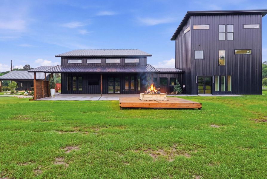 This modern home features a sleek black exterior with large windows for natural light. It includes a spacious backyard with a wooden deck and a cozy fire pit, perfect for outdoor gatherings.