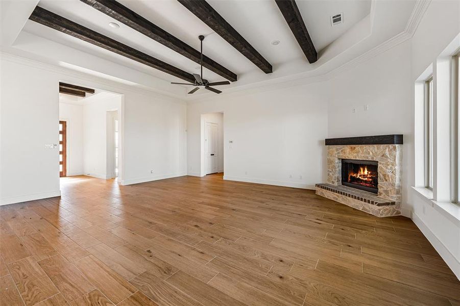 Unfurnished living room with a stone fireplace, light wood-type flooring, beam ceiling, ornamental molding, and a ceiling fan