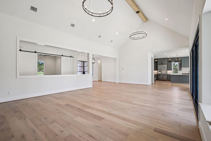 This spacious open-concept living area features high vaulted ceilings, modern light fixtures, and beautiful hardwood floors. It seamlessly connects to a sleek kitchen with stainless steel appliances and ample natural light from large windows and sliding glass doors. Perfect for entertaining and family living.