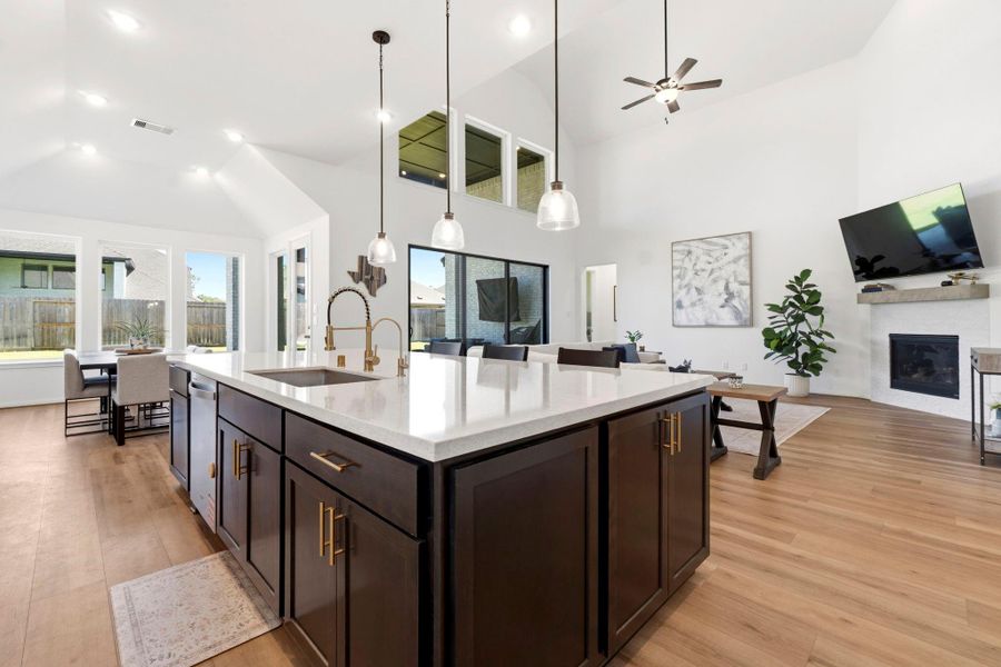 This open-concept living space features a modern kitchen with a large island, sleek pendant lighting, and ample cabinet storage. It flows into a bright dining area with large windows and a cozy living room with a fireplace. High ceilings and wood look vinyl flooring add to the spacious, contemporary feel.