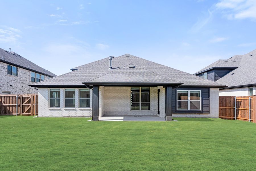 Rear exterior of single-story home with brick and dark siding, covered patio, and fenced backyard with green lawn Rear exterior of single-story home with brick and dark siding, covered patio, and fenced backyard with green lawn