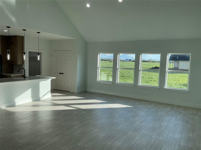 This photo shows a spacious, light-filled room with large windows overlooking a green landscape. The open layout includes a modern kitchen with a breakfast bar, pendant lighting, and light tile flooring, creating a bright and inviting atmosphere.
