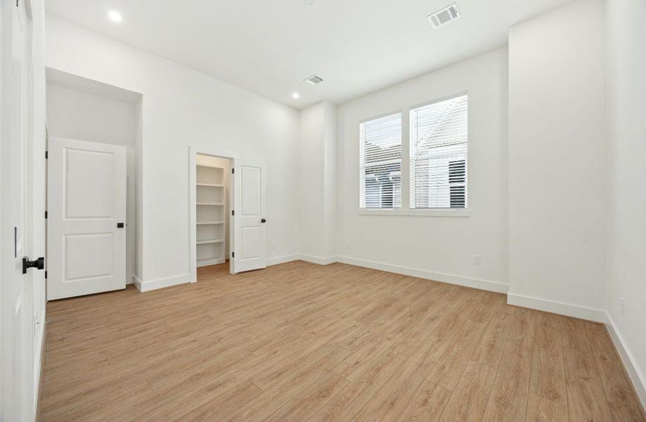Spacious upper-level bedroom with high ceilings, recessed lighting, warm wood-look flooring, large windows, and a walk-in closet with built-in shelving. The neutral palette and generous proportions create a calm, comfortable retreat ready for a variety of furniture layouts and personal styling.