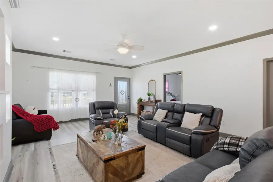 This spacious living room features modern gray sofas, a ceiling fan, and abundant natural light through large windows with sheer curtains. The room is accented with a wooden coffee table and light wood flooring, creating a cozy and inviting atmosphere.