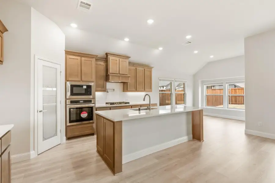 Kitchen with wood cabinets, center island with sink, double wall oven, and light hardwood floors open to living area Kitchen with wood cabinets, center island with sink, double wall oven, and light hardwood floors open to living area