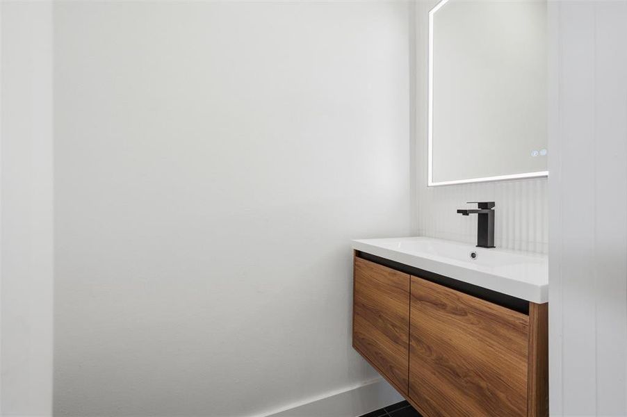 Secondary Bathroom features backlit vanity mirror positioned above a floating wood vanity with white countertop. Black faucet accents the space. Tile flooring extends throughout.