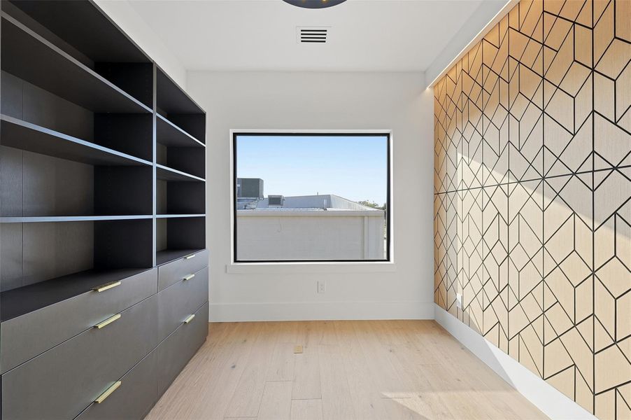Same floorplan - different finishes. Functional home office or dressing room showcasing a custom geometric wood-paneled accent wall and integrated dark-toned shelving with gold hardware.