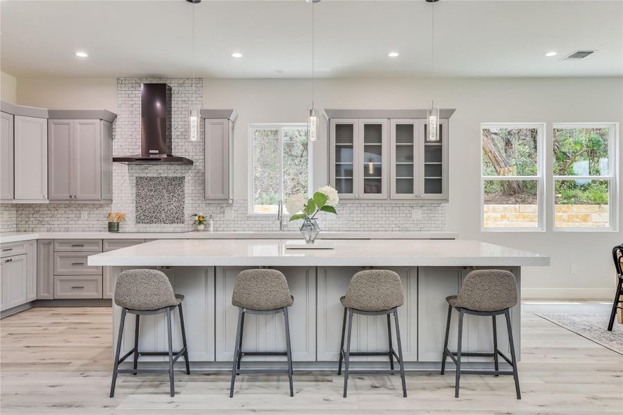 Kitchen featuring decorative light fixtures, wall chimney range hood, a healthy amount of sunlight, a kitchen island with sink, and a kitchen breakfast bar