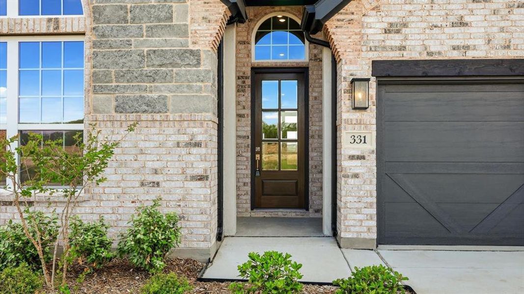 Two-tone brick and stone facade featuring an arched entryway with a dark wood-finish door and transom window, a paneled garage door, and modern exterior lighting