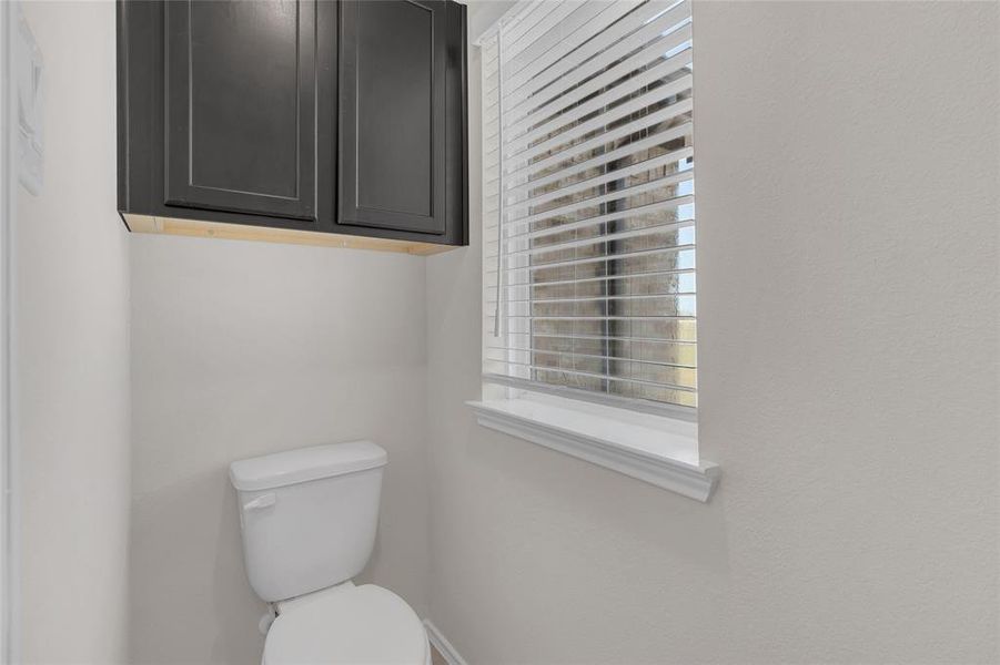 This is a compact bathroom featuring a white toilet and a dark cabinet overhead for storage, complemented by a window with white blinds, allowing for natural light.