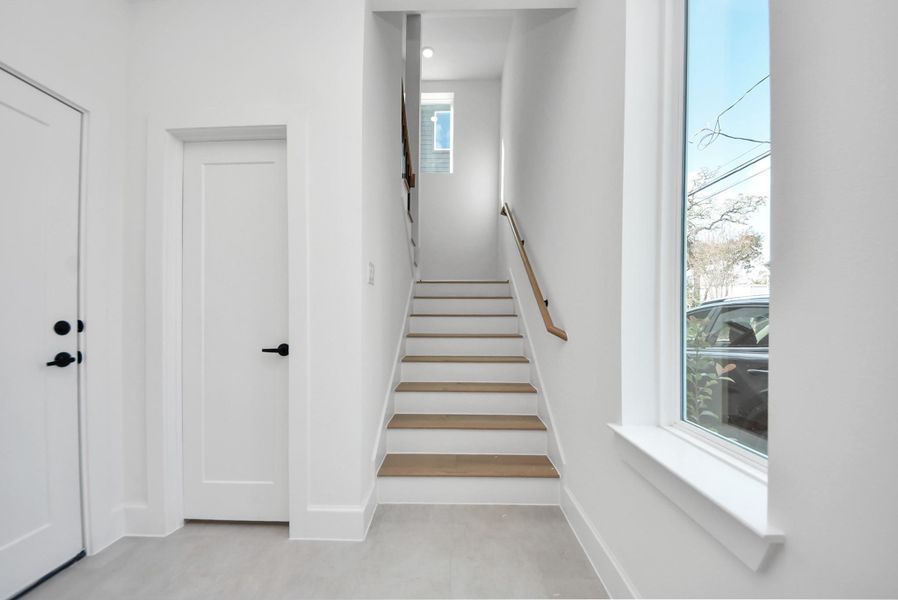 A bright, modern entryway with clean white walls, light flooring, and a staircase leading to the upper level. A large window brings in abundant natural light, while a simple handrail complements the contemporary design.