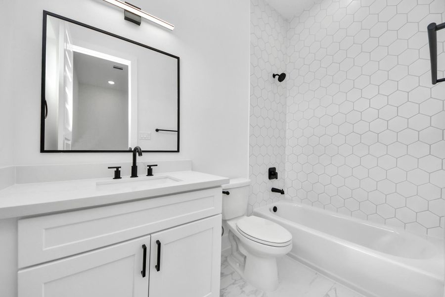 This bathroom features a modern design with sleek black fixtures, a large mirror, and a white vanity. The bathtub is surrounded by stylish hexagonal tiles, creating a clean and contemporary look.