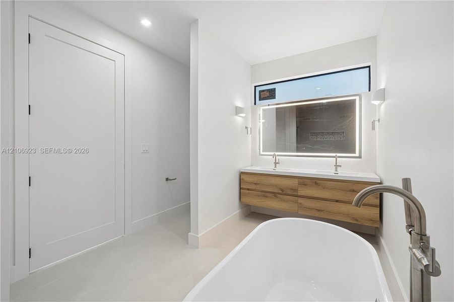 Freestanding soaking tub with floor-mounted brushed nickel faucet, floating wood-finish vanity with dual integrated sinks, and oversized LED-backlit mirror Freestanding soaking tub with floor-mounted brushed nickel faucet, floating wood-finish vanity with dual integrated sinks, and oversized LED-backlit mirror