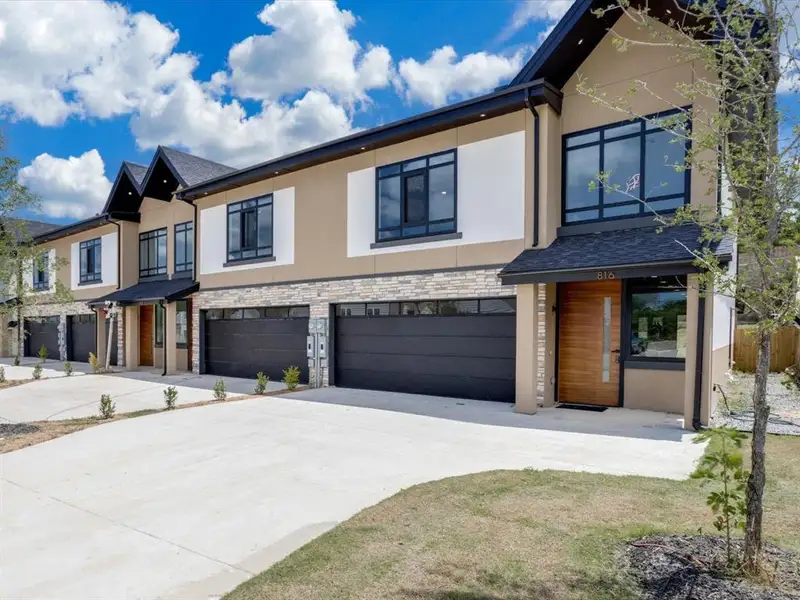 Modern townhome exterior featuring contemporary design, a striking wood-panel front door, black-framed windows, a stone-finish facade, and an attached garage Modern townhome exterior featuring contemporary design, a striking wood-panel front door, black-framed windows, a stone-finish facade, and an attached garage