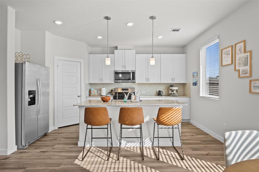 This modern kitchen features sleek white cabinetry, stainless steel appliances, and a central island with pendant lighting. The space is enhanced by natural light from a large window and offers seating with stylish barstools. A clean, open layout with wood flooring completes the inviting atmosphere.