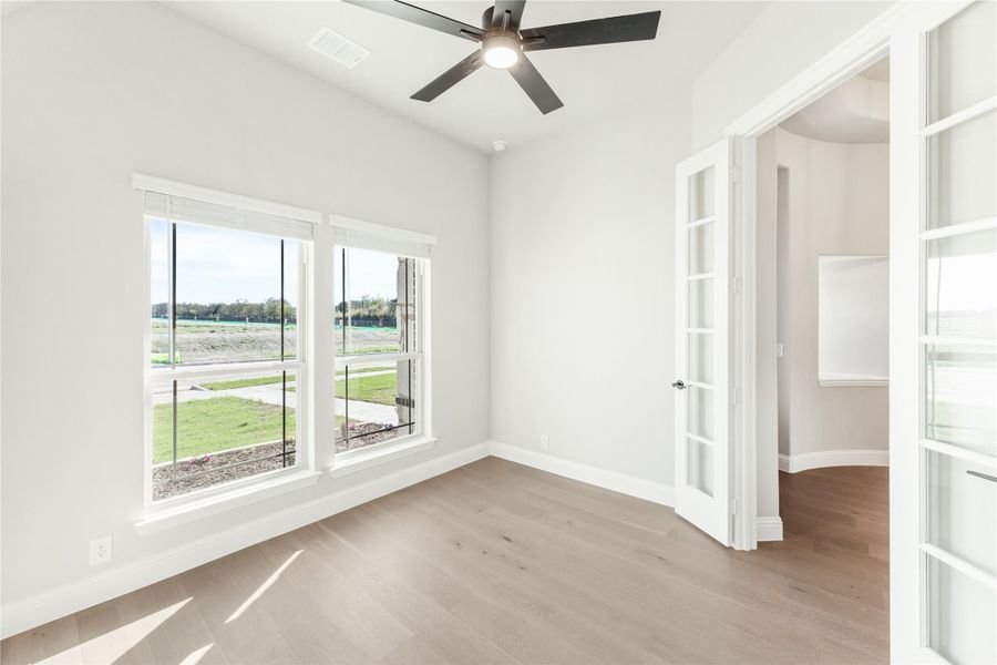 Empty room with hardwood floors, ceiling fan, large windows, and French doors with glass panels Empty room with hardwood floors, ceiling fan, large windows, and French doors with glass panels