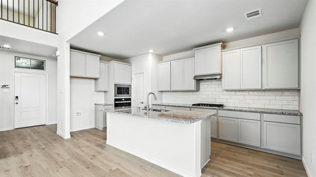 Contemporary kitchen featuring an island with a sink, stainless steel appliances, light gray cabinetry, subway tile backsplash, and wood-finish flooring