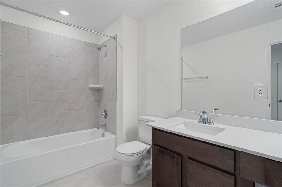 Secondary bath features tile flooring, bath/shower combo with tile surround, stained wood cabinets, beautiful light countertops, mirror, dark, sleek fixtures and modern finishes.