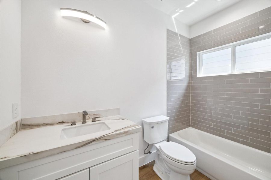 This full bathroom combines practical design with stylish finishes, featuring a marble-look vanity, under-mount sink, sleek faucet, and a tub/shower combination surrounded by glossy subway-style tile. A horizontal window introduces natural light while maintaining privacy. The bright neutral palette and clean-lined fixtures create a fresh, contemporary space for guests or household members.
