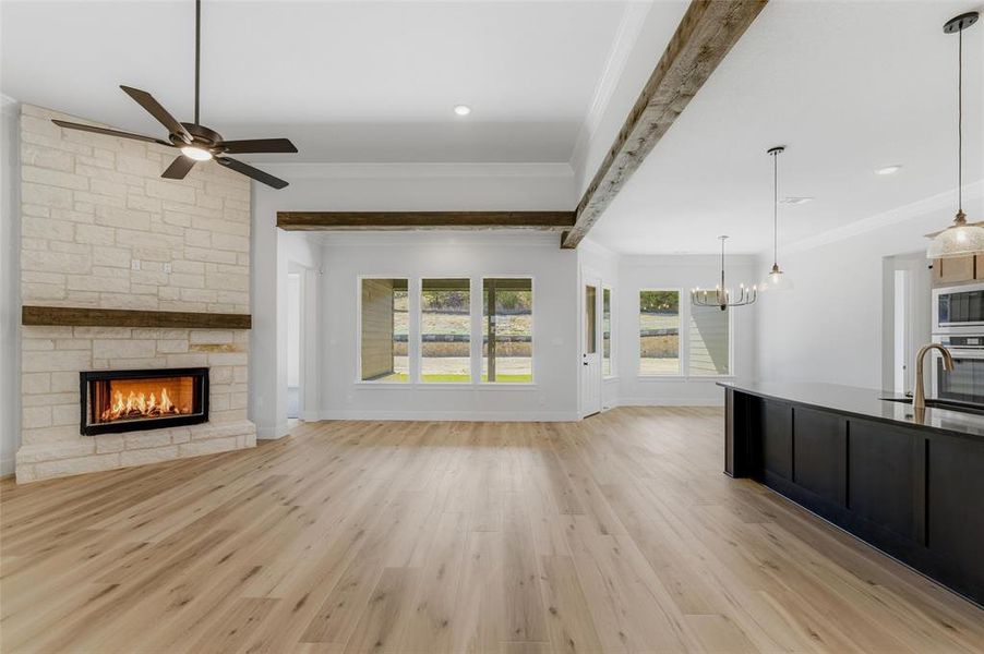 Unfurnished living room with light wood-style flooring, a stone fireplace, ornamental molding, beam ceiling, and ceiling fan