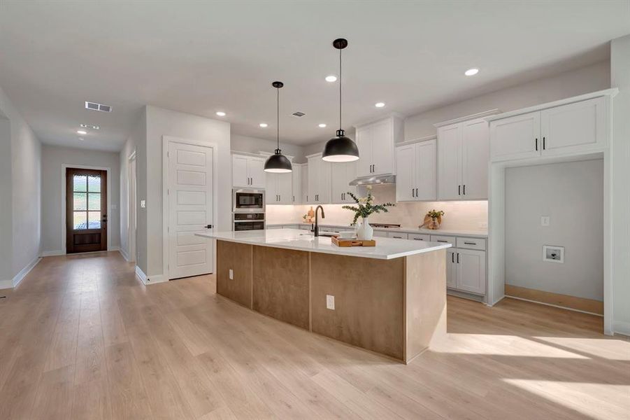 Kitchen with an island with sink, light wood-style floors, decorative light fixtures, stainless steel appliances, and light stone countertops